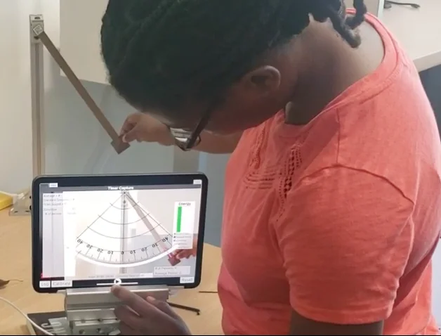 A student swinging a real pendulum while a tablet overlays an AR protractor and energy bar.